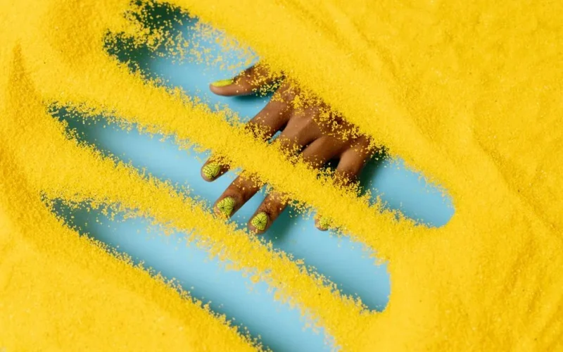 A striking image of a hand with yellow manicure submerged in vibrant yellow powder, showcasing creative nail art.