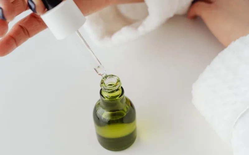 Close-up of hands with manicured nails holding a dropper over a green serum bottle.