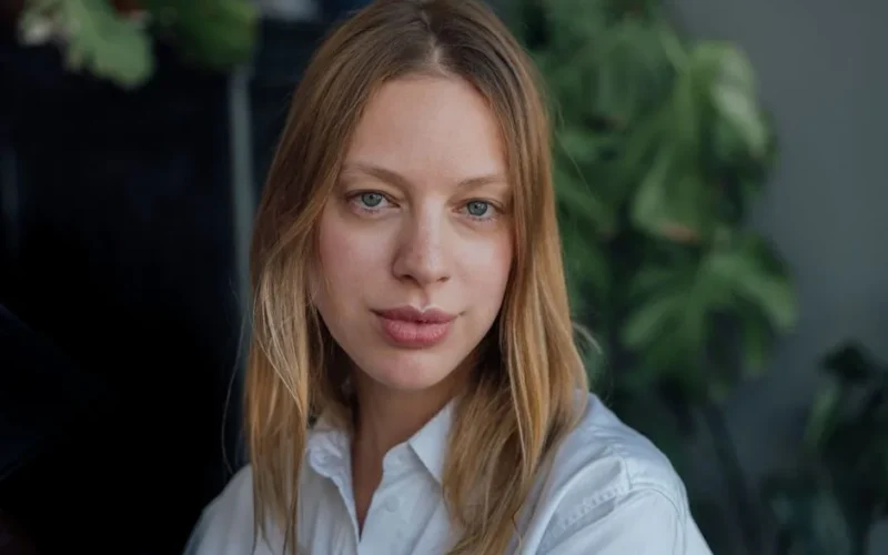 A close-up portrait of a woman with blonde hair and gray eyes in a white top.