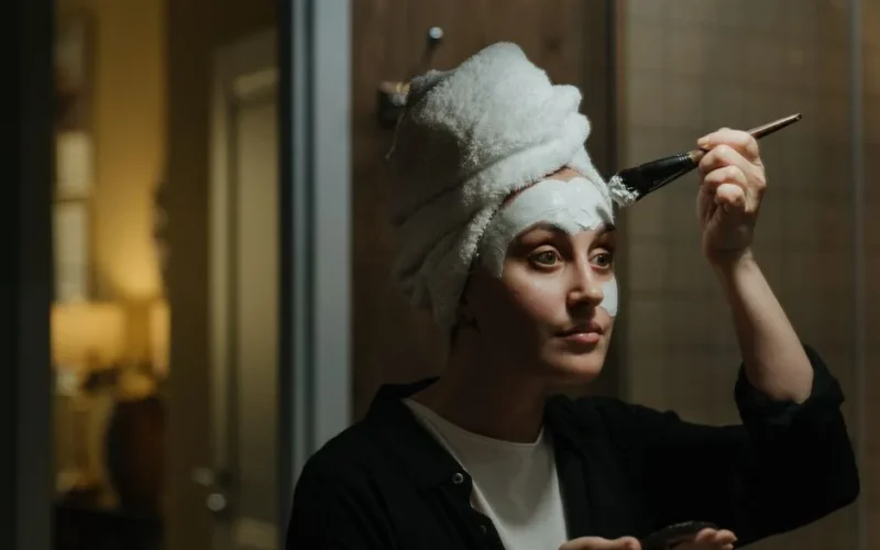 A woman applies a face mask in the bathroom with a towel on her head, emphasizing skincare and self-care.