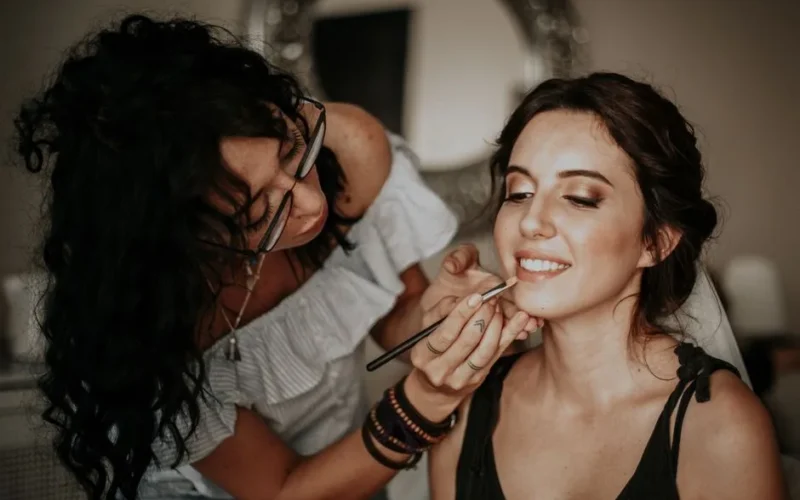 A makeup artist skillfully applies lipstick to a smiling woman in an indoor setting.