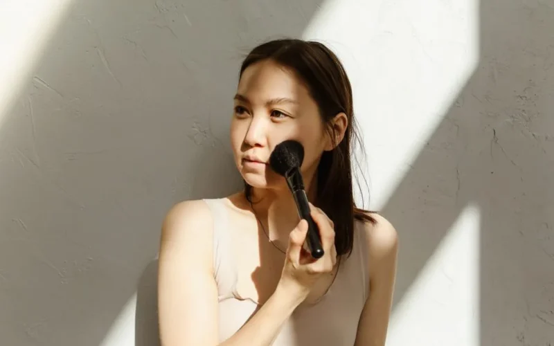 A young Asian woman applying makeup with a brush in natural lighting.