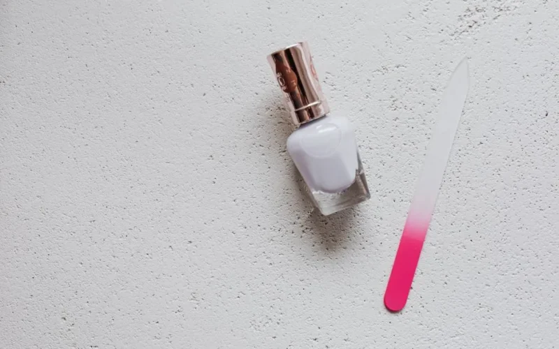 Flat lay of white nail polish and pink tinted glass nail file on a white textured background, ideal for beauty themes.