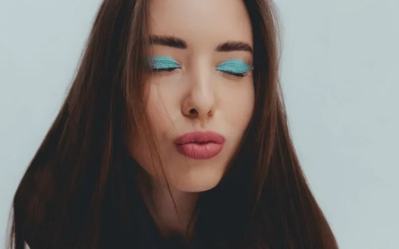 Studio portrait of a woman with vibrant blue eye shadow and pouting lips, eyes closed.