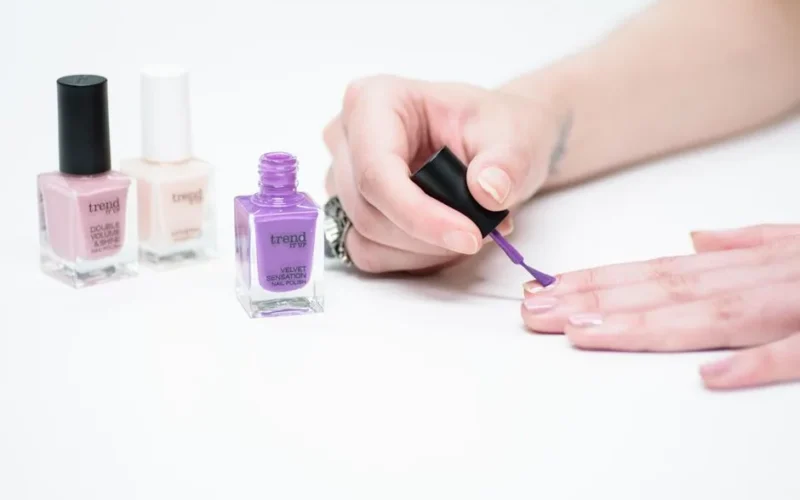 A woman applying purple nail polish with bottles of various colors in the background.