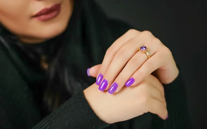 Close-up of woman's hands with vibrant purple nails and a gold ring, set against a dark background.