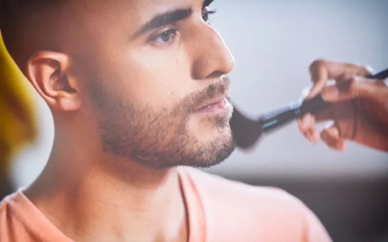 Close-up of a man having makeup applied with a brush by an artist.