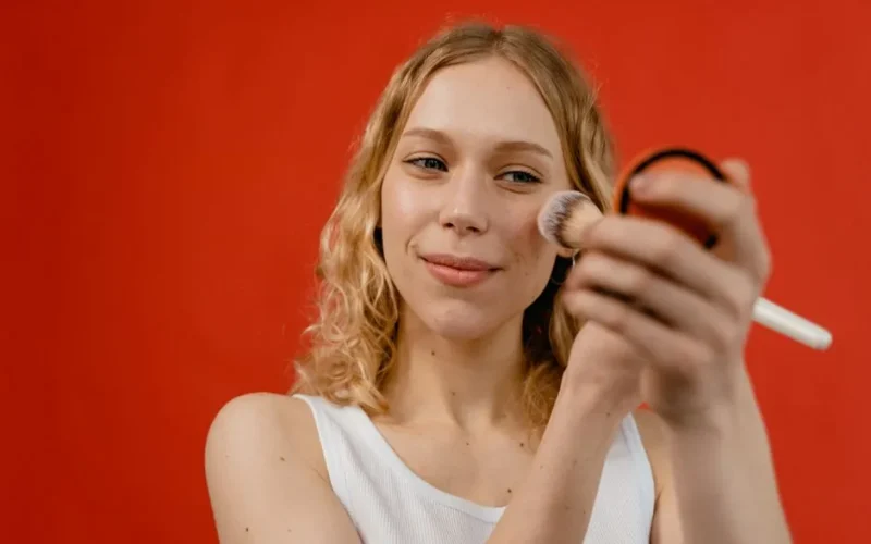 Woman applying makeup with a brush against a vibrant red backdrop, showcasing beauty and skincare concepts.