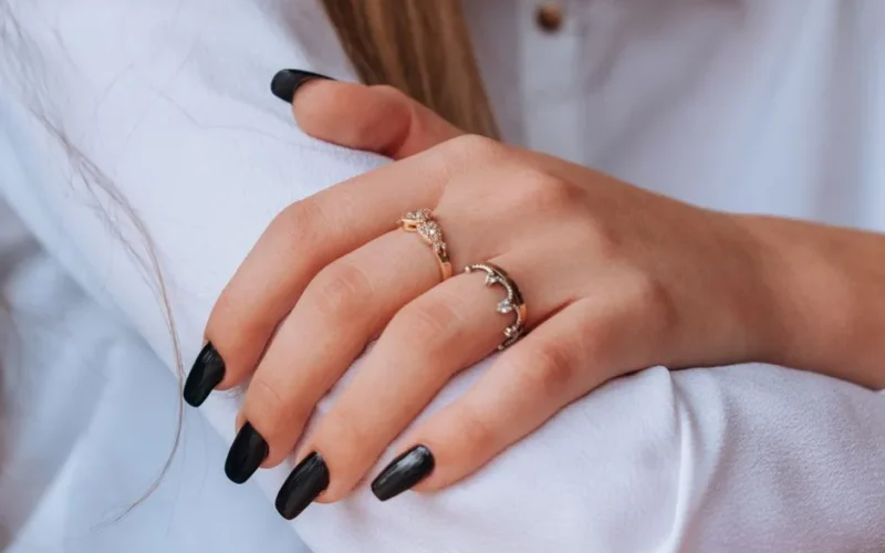 Close-up of hands with black nail polish and elegant rings against a white shirt backdrop.