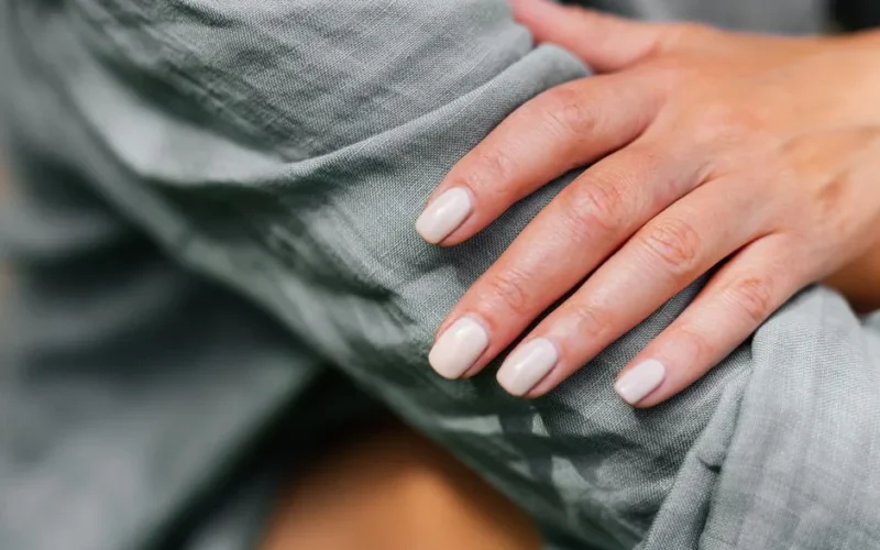 Close-up of a neatly manicured hand resting on a green sleeve, showcasing neutral nail polish.
