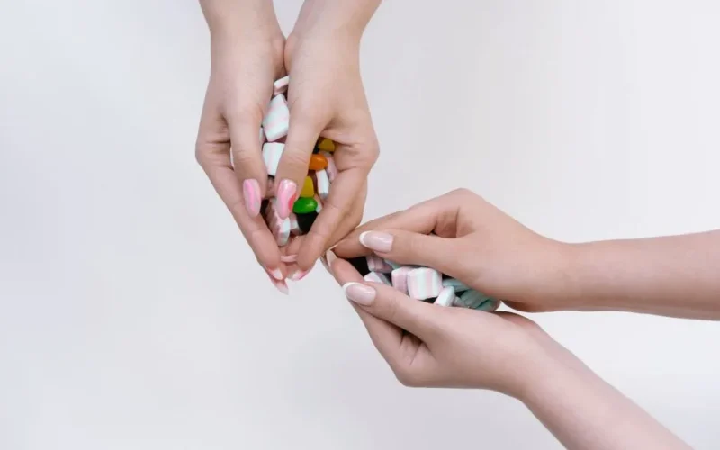 Three hands holding a mix of colorful candies on a white background, showcasing manicured nails.
