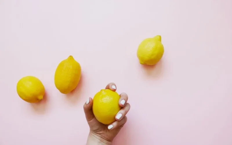 A hand with white nails holding a lemon on a soft pink background, surrounded by three additional lemons.