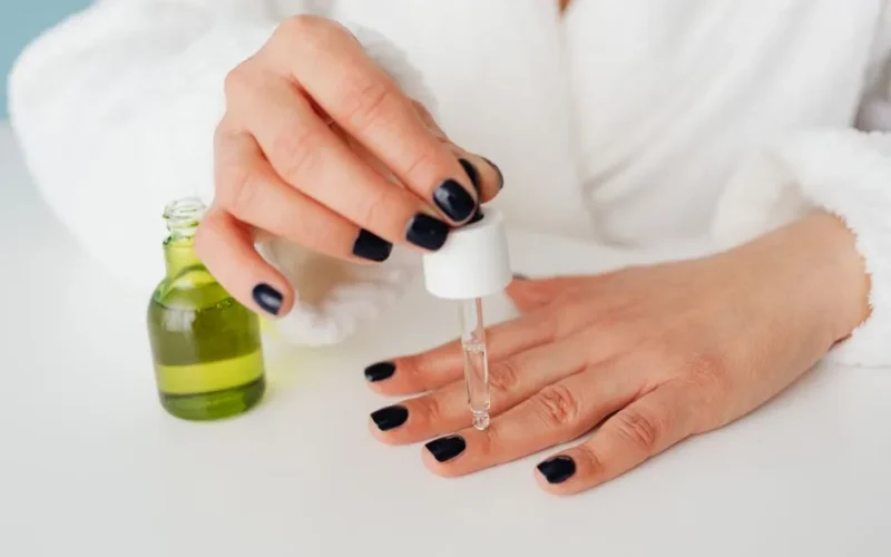 Close-up of a woman's hands applying serum to nails with a dropper in a bathrobe.