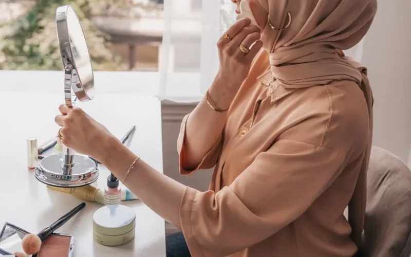 A woman in a hijab applies makeup at a bright indoor setting by a window.