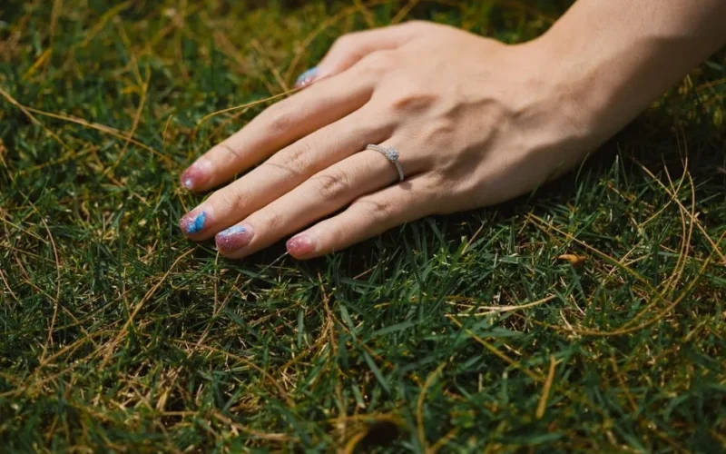 A hand with a ring and detailed manicure resting on green grass, showcasing elegance and beauty.