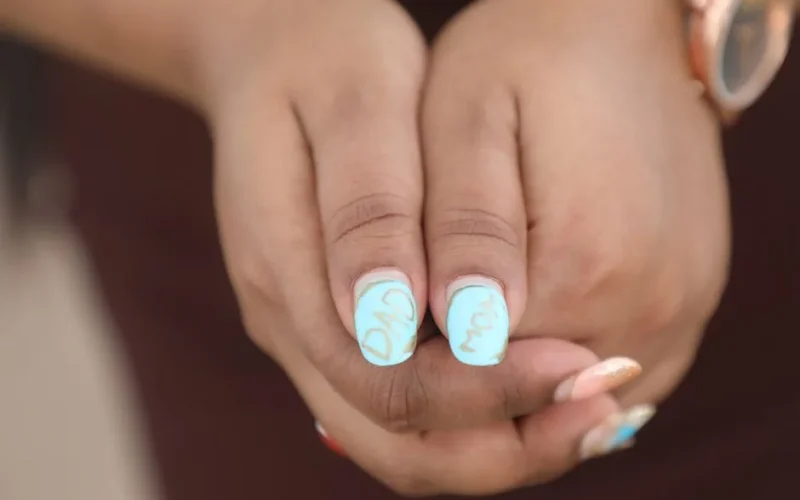 Detailed shot of hands featuring light blue nails with 'Mom' and 'Dad' written, showcasing personalized nail art.