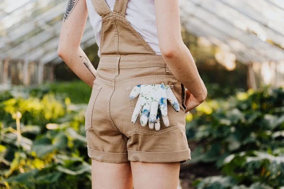 A woman in a garden wearing overalls with floral gloves in pocket, outdoor setting.
